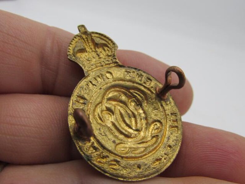 Puede incluir: Una insignia met&aacute;lica dorada, de forma circular, con una corona en la parte superior. La insignia presenta texto en relieve alrededor del borde y un dise&ntilde;o central. Dos presillas est&aacute;n unidas a la parte posterior para asegurar la insignia.