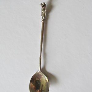 May include: Silver spoon with a detailed figurine of a person on the handle. The spoon has a rounded oval bowl.