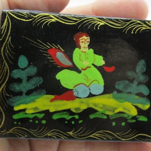 May include: A small, rectangular black box with a painted scene. The scene depicts a figure in green clothing riding a red object, with green trees and yellow ground. The box has a gold decorative border.