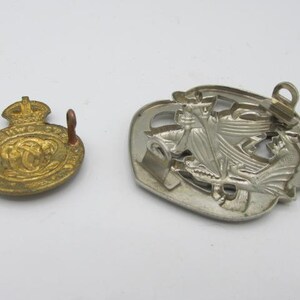 Puede incluir: Dos insignias met&aacute;licas vintage. Una es dorada, circular y presenta una corona y texto. La otra es plateada, en forma de escudo y representa un emblema detallado. Ambas insignias tienen alfileres.