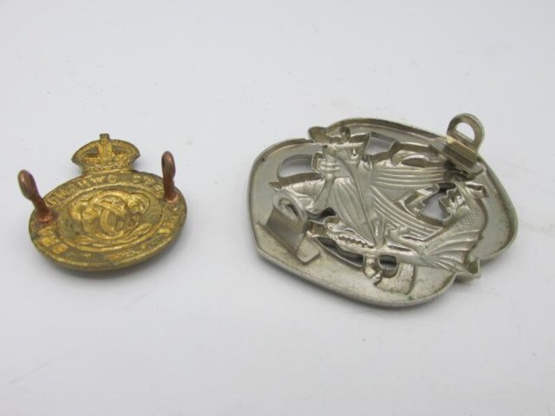 Puede incluir: Dos insignias met&aacute;licas vintage. Una es dorada, circular y presenta una corona y texto. La otra es plateada, en forma de escudo y representa un emblema detallado. Ambas insignias tienen alfileres.