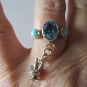 Puede incluir: Anillo de plata con una gran piedra azul ovalada, dos piedras turquesas más pequeñas y un dije de jarrón en miniatura. El anillo se lleva en un dedo, mostrando el diseño intrincado y el contraste de colores.