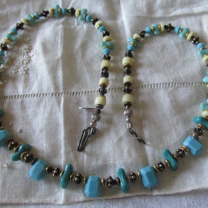 May include: A turquoise and cream-colored beaded necklace with gold-toned accents. The necklace features a variety of bead shapes and sizes, including faceted turquoise beads and cream-colored cylindrical beads. The necklace is displayed on a white, textured surface.