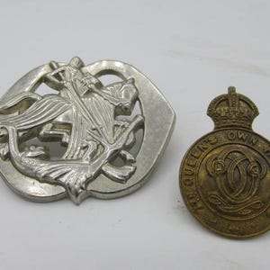 Puede incluir: Dos insignias met&aacute;licas antiguas. Una es plateada, con una figura a caballo, y la otra es una insignia m&aacute;s peque&ntilde;a, dorada, con el texto "QUEEN'S OWN HUSSARS" alrededor de un dise&ntilde;o central.