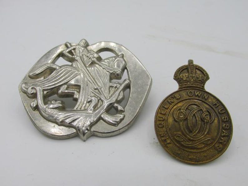 Puede incluir: Dos insignias met&aacute;licas antiguas. Una es plateada, con una figura a caballo, y la otra es una insignia m&aacute;s peque&ntilde;a, dorada, con el texto "QUEEN'S OWN HUSSARS" alrededor de un dise&ntilde;o central.