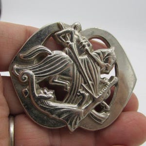 Puede incluir: Broche plateado con un relieve detallado de un caballero a caballo matando a un drag&oacute;n. La broche tiene una parte superior arqueada y un dise&ntilde;o circular, con detalles intrincados.