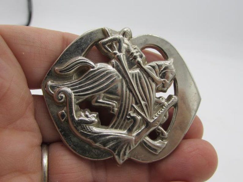 Puede incluir: Broche plateado con un relieve detallado de un caballero a caballo matando a un drag&oacute;n. La broche tiene una parte superior arqueada y un dise&ntilde;o circular, con detalles intrincados.