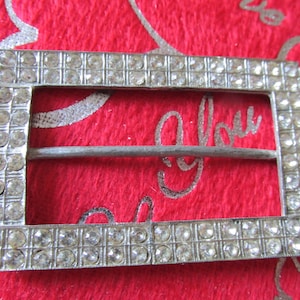May include: A rectangular silver-toned buckle encrusted with small, clear rhinestones. The buckle has a central bar and is set against a red background with the word "You" in cursive script.
