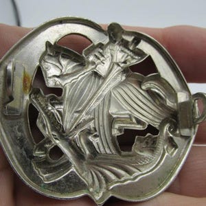 Puede incluir: Insignia de metal plateado que representa a un caballero a caballo matando a un drag&oacute;n. El relieve detallado muestra al caballero con una espada, el caballo y el drag&oacute;n. La insignia tiene un borde decorativo y un cierre en la parte posterior.