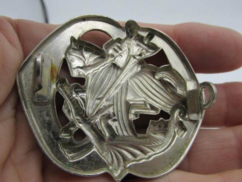 Puede incluir: Insignia de metal plateado que representa a un caballero a caballo matando a un drag&oacute;n. El relieve detallado muestra al caballero con una espada, el caballo y el drag&oacute;n. La insignia tiene un borde decorativo y un cierre en la parte posterior.