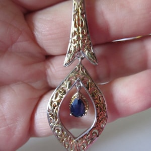 Op de afbeelding: Een zilveren en gouden hanger met een blauwe edelsteen in het midden. De hanger heeft de vorm van een diamant met ingewikkelde filigraan details.