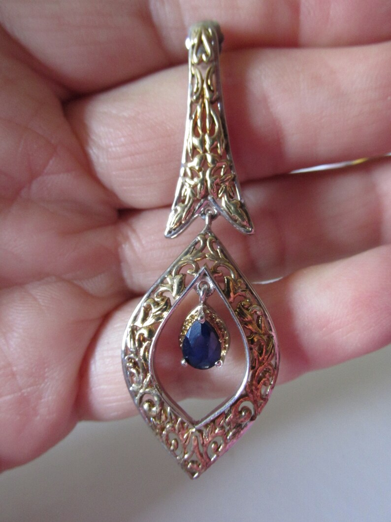 Op de afbeelding: Een zilveren en gouden hanger met een blauwe edelsteen in het midden. De hanger heeft de vorm van een diamant met ingewikkelde filigraan details.