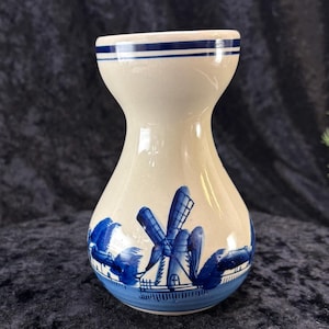 May include: A white ceramic vase with a blue and white windmill design. The vase has a wide mouth and a narrow neck.