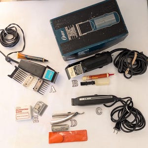May include: A collection of vintage Oster electric hair clippers and accessories, including a box, blades, and a small bottle of oil. The clippers are black and silver with a blue and white logo. The box is blue and black with the Oster logo and the words "Detachable Blade Clipper".