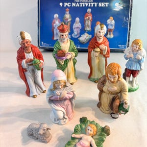 May include: A 9-piece fine porcelain nativity set featuring hand-painted figurines including the infant in a manger, a kneeling figure, a lamb, and various standing figures in robes. The set includes a blue box labeled 9 PC NATIVITY SET.