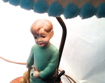 May include: A vintage lamp with a blue and white polka dot shade and a blue pom-pom trim. The lamp features a figurine of a boy holding a dog and a set of colorful blocks.
