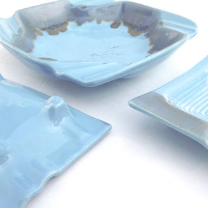 Könnte beinhalten: Drei hellblaue Keramik-Aschenbecher mit quadratischer Form und gewellten Kanten. Die Aschenbecher haben eine glänzende Oberfläche und ein dekoratives Design im Inneren. Die Ränder sind mit einer dunkleren blau-grauen Farbe akzentuiert.