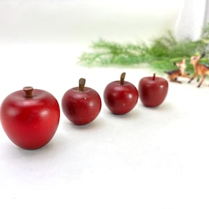 May include: Four red decorative apples with brown stems are arranged in a row. The apples have a glossy finish and are set against a white background. Miniature deer figurines and greenery are visible in the background.