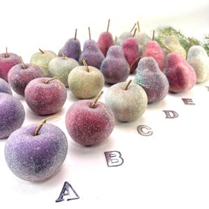 May include: A collection of decorative, glitter-covered apples and pears in various colors, including red, purple, and green. Each piece has a small stem and a gold string for hanging. The fruits are arranged on a white surface.
