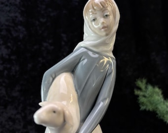 May include: A porcelain figurine of a girl wearing a blue dress and a white headscarf, holding a white lamb in her arms.