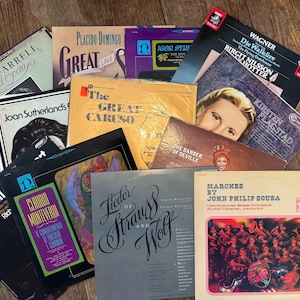 May include: A collection of vintage vinyl records featuring opera singers like Eileen Farrell, Joan Sutherland, and Placido Domingo. The records are arranged in a stack on a wooden surface. Some of the record covers feature colorful artwork and text.