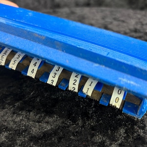 Könnte beinhalten: Ein blauer Plastik-Zählwerk mit weißen Etiketten, die von 0 bis 6 nummeriert sind. Der Zähler wird zum Zählen von Gegenständen oder zum Verfolgen von Zahlen verwendet.