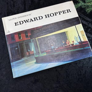 Peut inclure: Un livre intitulé "Edward Hopper" de Lloyd Goodrich, présentant une peinture d'un restaurant avec des personnes assises au comptoir. Le restaurant s'appelle "The Phillies" et a une marquise jaune.