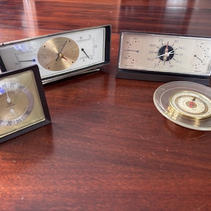 May include: Four vintage weather instruments on a wooden surface. The collection includes two rectangular barometers, a square barometer, and a small, round thermometer. The barometers have gold and silver faces with black frames. The thermometer is in a clear, round case.
