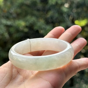 May include: A white jade bangle bracelet with a smooth, polished finish. The bracelet is held in a hand, with a blurred green background.