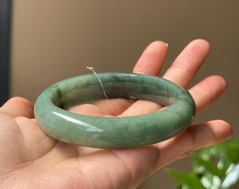 May include: A green jade bangle bracelet. The bangle is smooth and polished, with a slightly rounded shape.