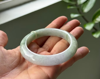 May include: A light green and white jade bangle bracelet. The smooth, polished surface reflects light, showcasing the natural variations in color. The bracelet is a circular shape, designed to be worn on the wrist. A delicate, light green string is wrapped around the bracelet.