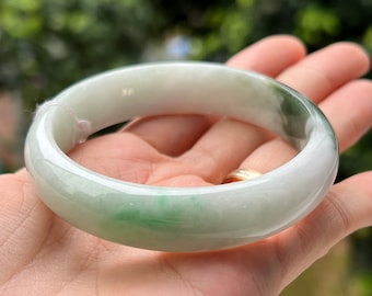 May include: A jade bangle bracelet with a light green colour and white swirls. The bangle is smooth and polished.