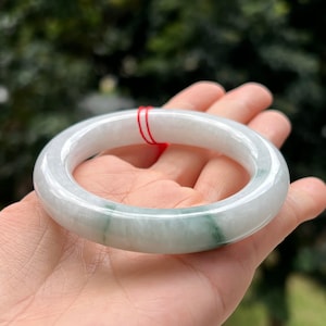 Könnte beinhalten: Ein Jade-Armband mit hellgrüner Farbe und weißen Streifen. Das Armband ist glatt und poliert und mit einer roten Schnur gebunden.