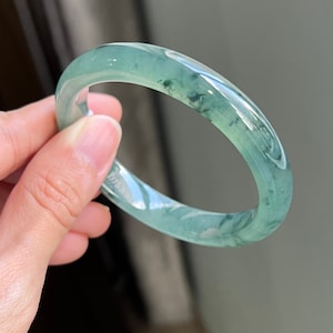 May include: A jade bangle bracelet with a light green color and a swirling pattern. The bangle is smooth and polished.