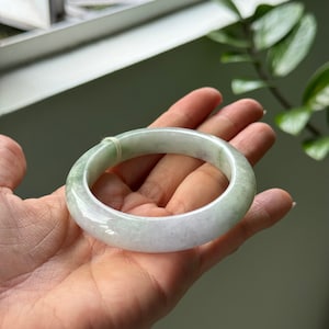May include: A light green and white jade bangle bracelet. The smooth, polished surface reflects light, showcasing the natural variations in color. The bracelet is a circular shape, designed to be worn on the wrist. A delicate, light green string is wrapped around the bracelet.