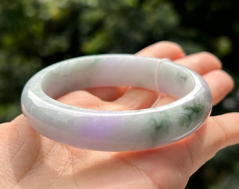 May include: A smooth, polished bangle bracelet crafted from translucent jadeite stone. The bangle features a rounded profile with a pale lavender base color, accented by distinct patches of deep forest green and soft white marbling throughout.