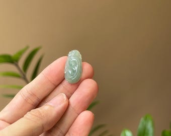 Puede incluir: Un colgante de jade verde claro tallado con un diseño de espiral. El colgante tiene forma ovalada y un acabado liso y pulido.