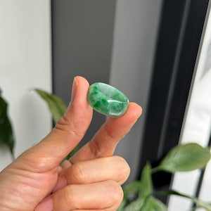 Puede incluir: Un vibrante anillo de jade verde con una superficie pulida y lisa. El anillo se sostiene entre dos dedos, mostrando sus variaciones de color naturales. El anillo de jade es una pieza de joyería clásica.