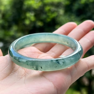 May include: A jade bangle bracelet with a light green color and dark green inclusions. The bangle is smooth and polished.
