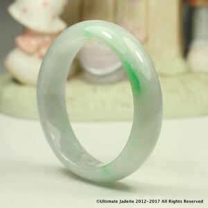 Può includere: Un bracciale bangle in giada traslucida, con una miscela di tonalità verde pallido e bianco. La superficie lucida riflette la luce, mettendo in risalto le variazioni naturali di colore. Un gioiello classico.