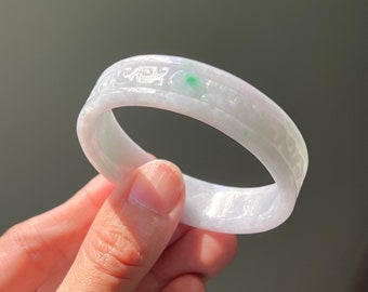 May include: A translucent white jade bangle bracelet featuring a smooth, polished surface with intricate carved patterns along the outer band and a single, small, vibrant green spot embedded within the stone's natural, cloudy, and pale lavender texture.