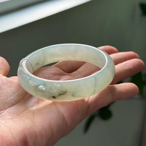 Può includere: Un braccialetto di giada verde chiaro con una finitura liscia e lucida. Il braccialetto è tenuto in una mano, mostrando le sue dimensioni e la sua forma.
