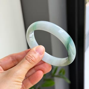 Può includere: Un braccialetto bangle in giada traslucida. Il braccialetto presenta una superficie liscia e lucida con una miscela di tonalità verde pallido e bianco. La giada è tenuta in una mano, mostrando le sue variazioni di colore naturali e il suo design elegante.