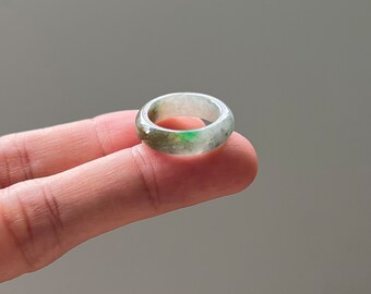 May include: A jade ring with a smooth, rounded design. The ring features a mix of green and white hues, with a translucent quality. The ring is held by a finger, showcasing its size and shape. A piece of jewelry.