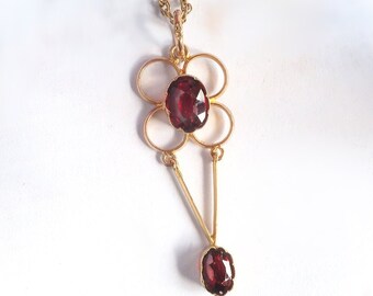 May include: A gold pendant necklace with a delicate design featuring two red gemstones. The pendant is shaped like a flower with four rounded petals and a central gemstone. A second gemstone hangs from the bottom of the pendant.