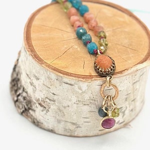 Puede incluir: Un collar de múltiples hilos con cuentas azules, verdes y naranjas. El collar tiene un colgante con una piedra naranja tallada y un dije con cuentas más pequeñas.