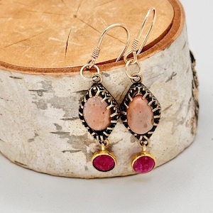 May include: A pair of dangle earrings featuring a pink oval stone set in a gold-toned filigree frame. Each earring has a small pink gemstone hanging below the stone.
