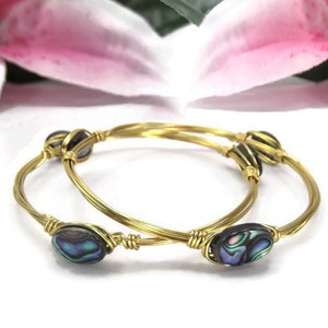 Puede incluir: Dos pulseras de alambre dorado envueltas con acentos de concha de abulón iridiscente.