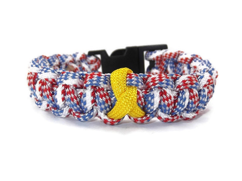 May include: A braided paracord bracelet in red, white, and blue with a black buckle. A yellow awareness ribbon is centered on the bracelet. The bracelet is a patriotic accessory.