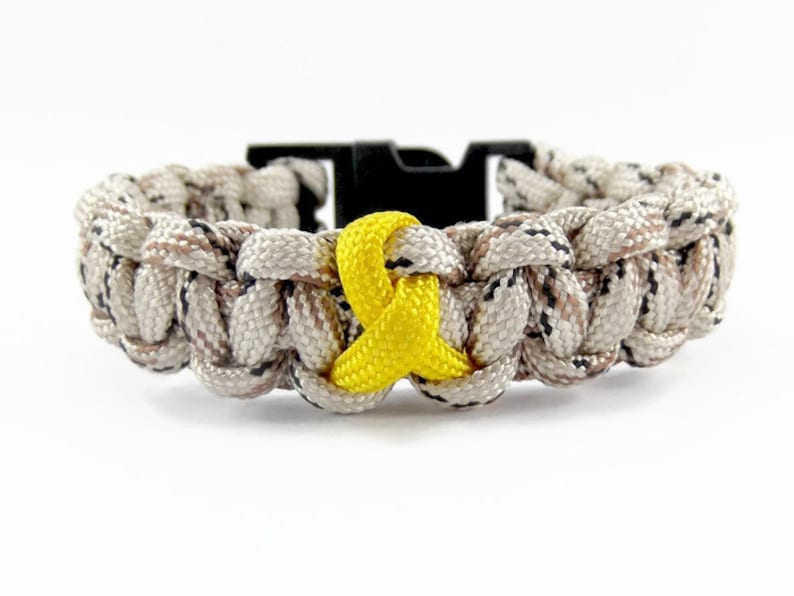 May include: A camouflage paracord bracelet with a yellow ribbon charm. The bracelet is made of braided cord and has a black plastic buckle.